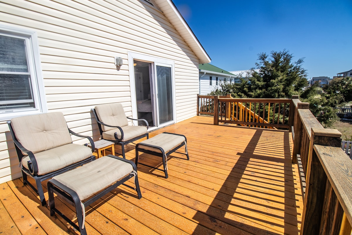 A wooden deck features two lounge chairs and a small table, inviting relaxation outdoors. The sun casts distinct shadows on the floorboards, enhancing the spacious area. Surrounding greenery and adjacent buildings provide a sense of privacy while ensuring a bright, open atmosphere.