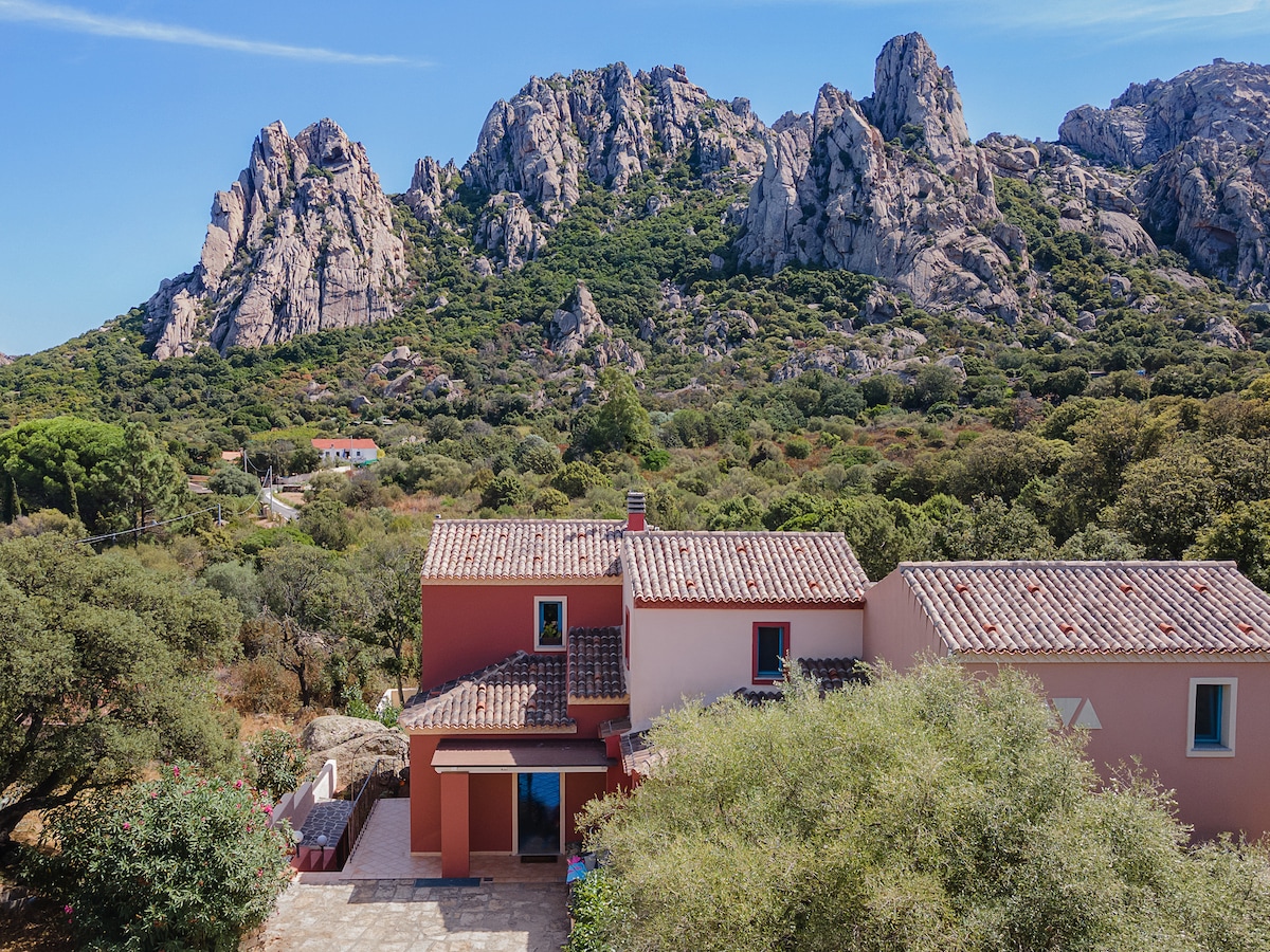 A charming three-story house is nestled among lush greenery, with rocky mountains rising in the background. The earthy colors of the exterior blend harmoniously with the natural surroundings, while ample outdoor space is present for relaxation.