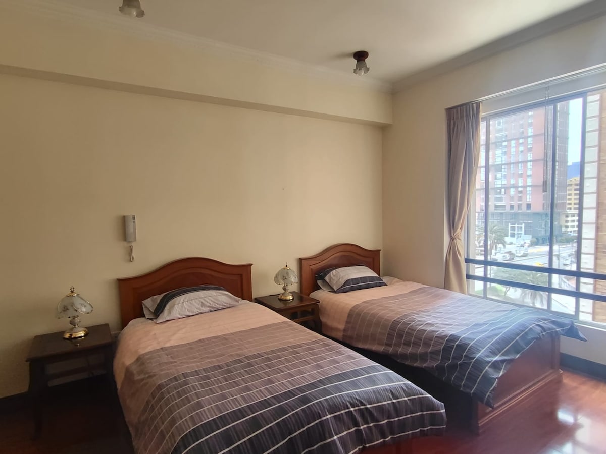 A well-lit room features two wooden beds with coordinated bedding, situated beside small side tables with lamps. A large window offers a view of the outside, with natural light illuminating the space and highlighting the warm tones of the wooden furniture.