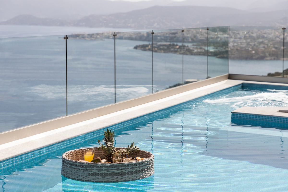 A serene swimming pool extends to the edge, offering panoramic views of the bay and landscape. A floating tray features refreshing drinks and fruit, enhancing the tranquil ambiance. Glass railings provide unobstructed views, ensuring a seamless connection to the stunning surroundings.