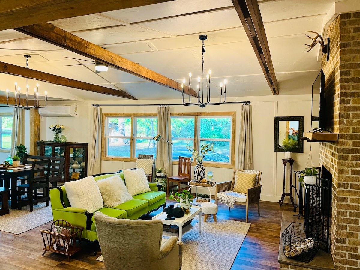 A cozy living area features a mix of comfortable seating with green and neutral tones. Large windows allow natural light, revealing views of the surrounding nature. A rustic coffee table sits in the center, and the room is accented by wooden beams overhead.