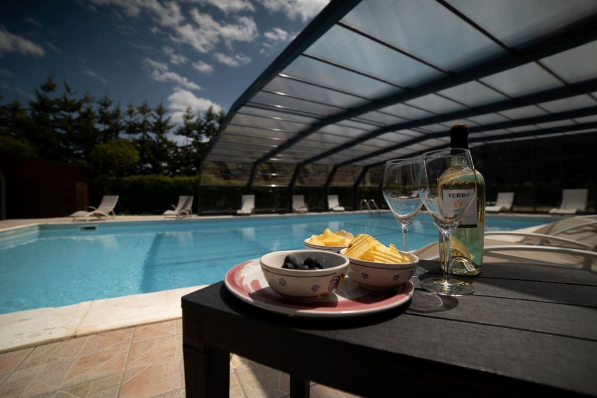 A well-appointed table is set near the pool, featuring a bottle of wine and two glasses alongside a plate with olives and chips. The pool, covered for protection, reflects the blue sky and surrounding greenery, with comfortable loungers positioned for relaxation.