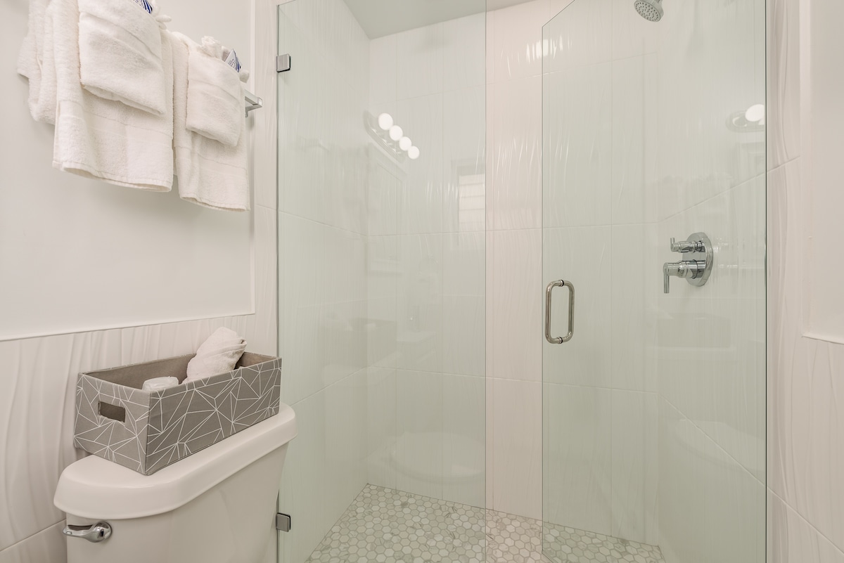 A modern bathroom setting is displayed, featuring a glass shower with a rainfall showerhead. Light tiles reflect a clean aesthetic, complemented by a geometric storage basket holding rolled towels. Soft lighting is provided by round fixtures above the mirror, creating a light and airy feel.