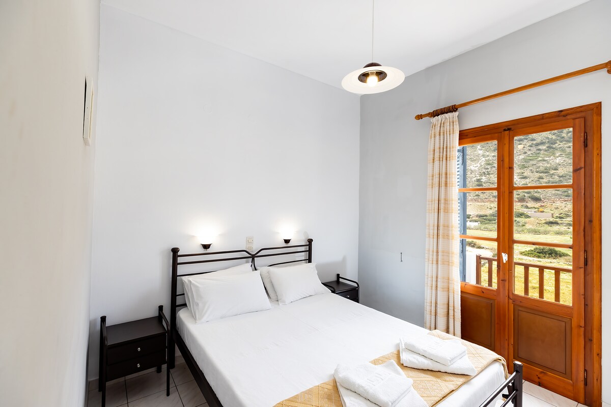 A bright bedroom is equipped with a spacious bed made with fresh linens. Two nightstands are positioned on either side of the bed. Natural light enters through a pair of wooden doors that lead to a balcony, with scenic views of the surrounding hills.