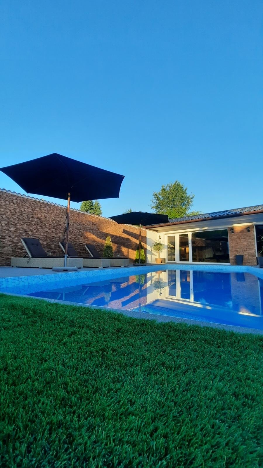 A private outdoor area features a swimming pool with a clear blue surface, surrounded by manicured grass. Sun loungers are positioned beside the pool, while a large umbrella provides shade. The setting is enhanced by a backdrop of a brick wall and the edge of the house.
