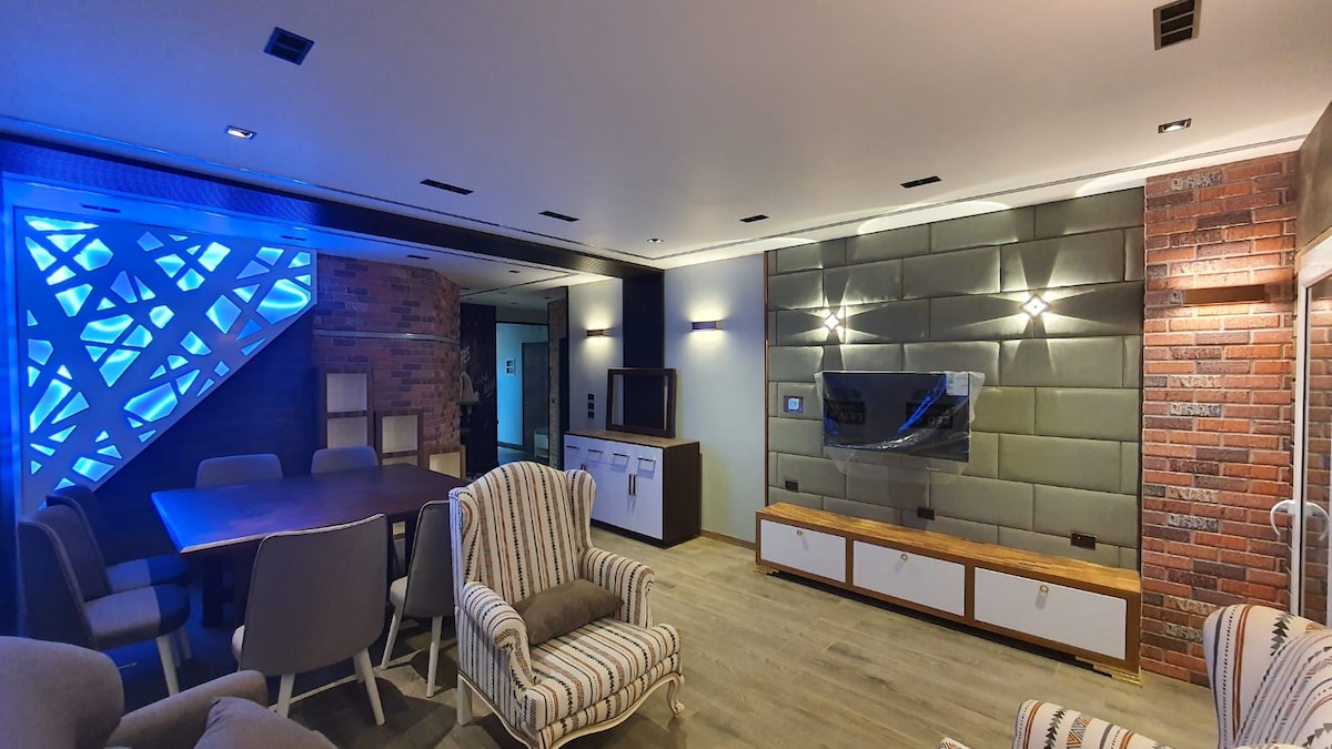 A modern living area features a mix of textured walls and contemporary furniture. A spacious table is surrounded by gray chairs, and a soft, striped armchair adds comfort. The area is illuminated by recessed lighting and a blue-lit accent wall, creating a distinctive ambiance.