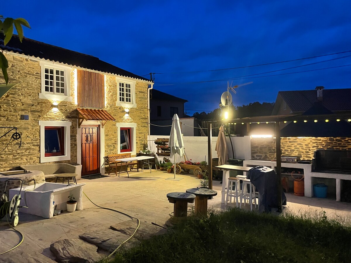 The exterior of the house is illuminated at night, highlighting its stone façade and warm wooden accents. A spacious outdoor area features seating arrangements, umbrellas, and a barbecue setup. Decorative lighting strands create a welcoming ambiance for evening gatherings.