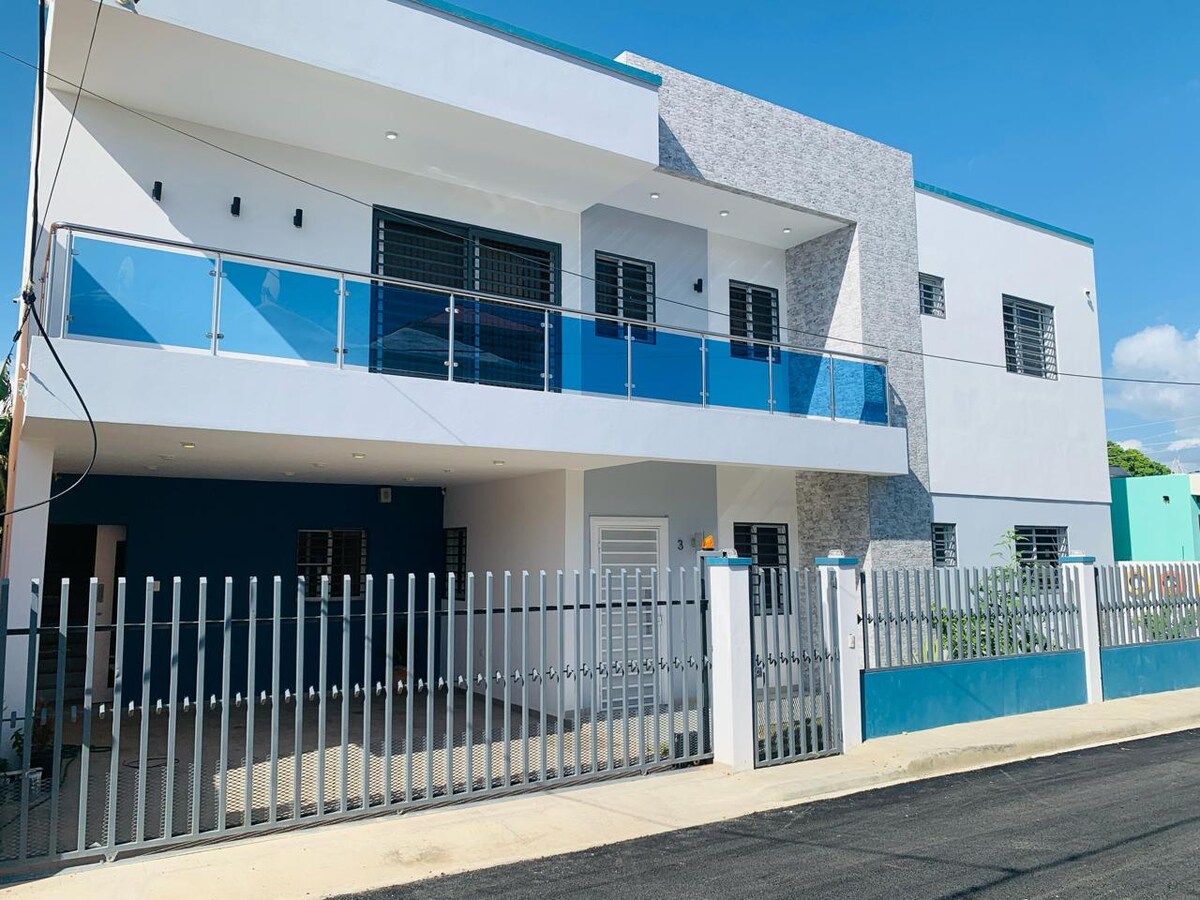 A modern two-story building is presented, featuring a contemporary façade with large windows and balconies. Glass railings accentuate the exterior space, while a gated entrance is visible. The vibrant blue sky complements the clean white walls, contributing to a refreshing aesthetic.