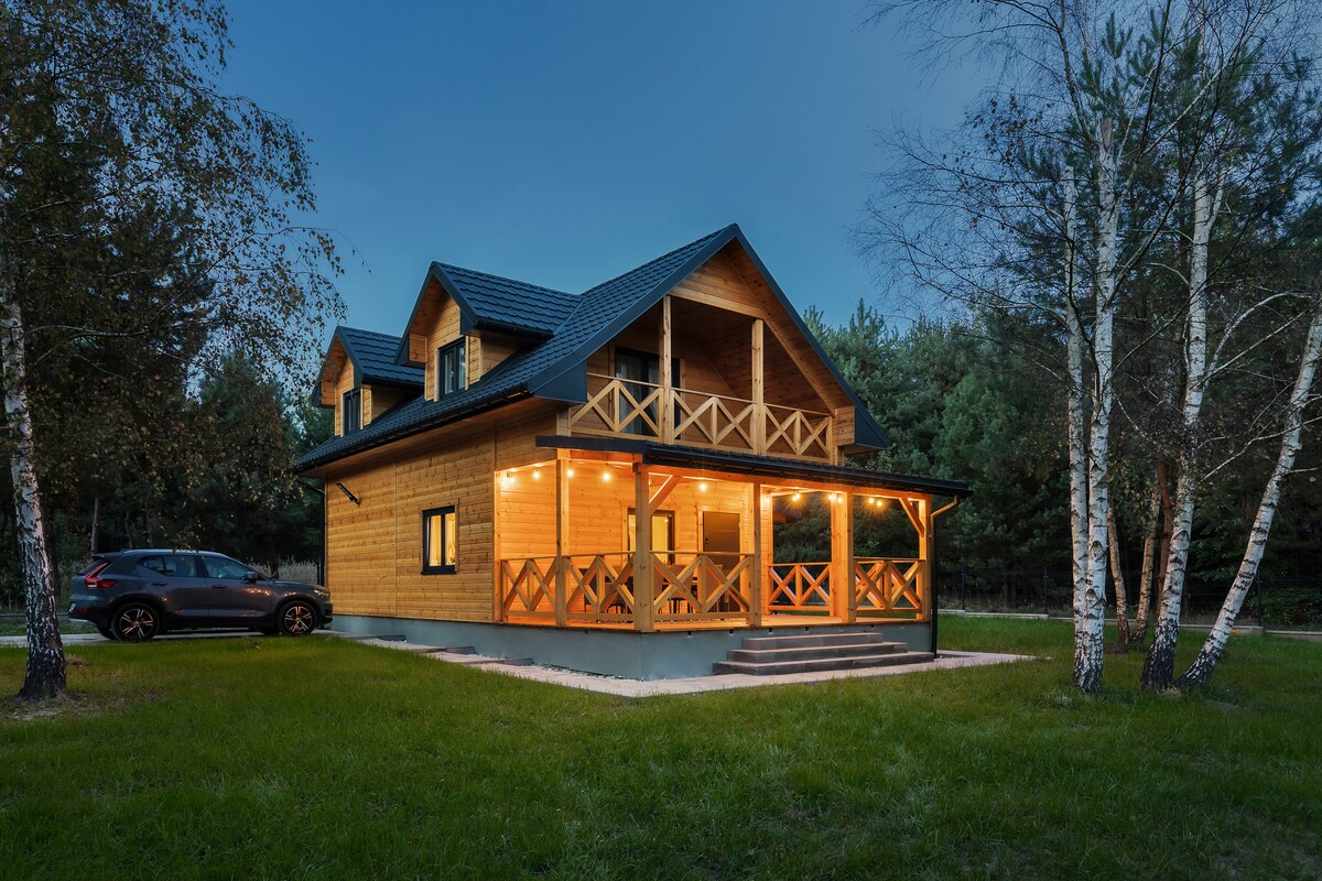A wooden cabin is showcased, featuring a spacious covered terrace with wooden railings. Soft lighting highlights the house's architectural details. The surrounding green grass and trees, along with a parked car nearby, enhance the natural setting.
