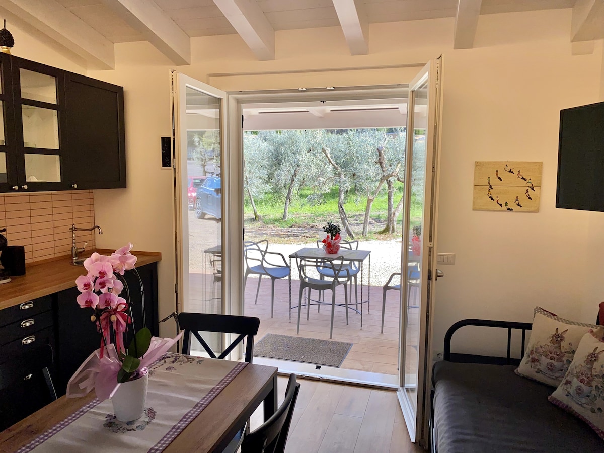 Large glass doors open to a patio area surrounded by olive trees. A wooden table and chairs are placed outside, complemented by a potted flower arrangement. The interior showcases a cozy seating area and a kitchen space with black cabinetry and light wood accents.