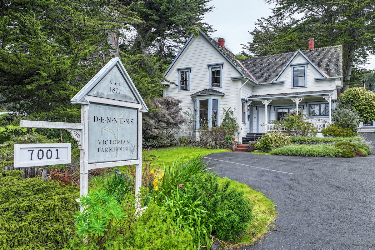 The exterior of Dennen's Victorian Farmhouse showcases a charming two-story structure, framed by lush greenery and colorful flowering plants. A welcoming sign displays the establishment's name and year established. The driveway leads to the entrance, enhancing the property's inviting presence.