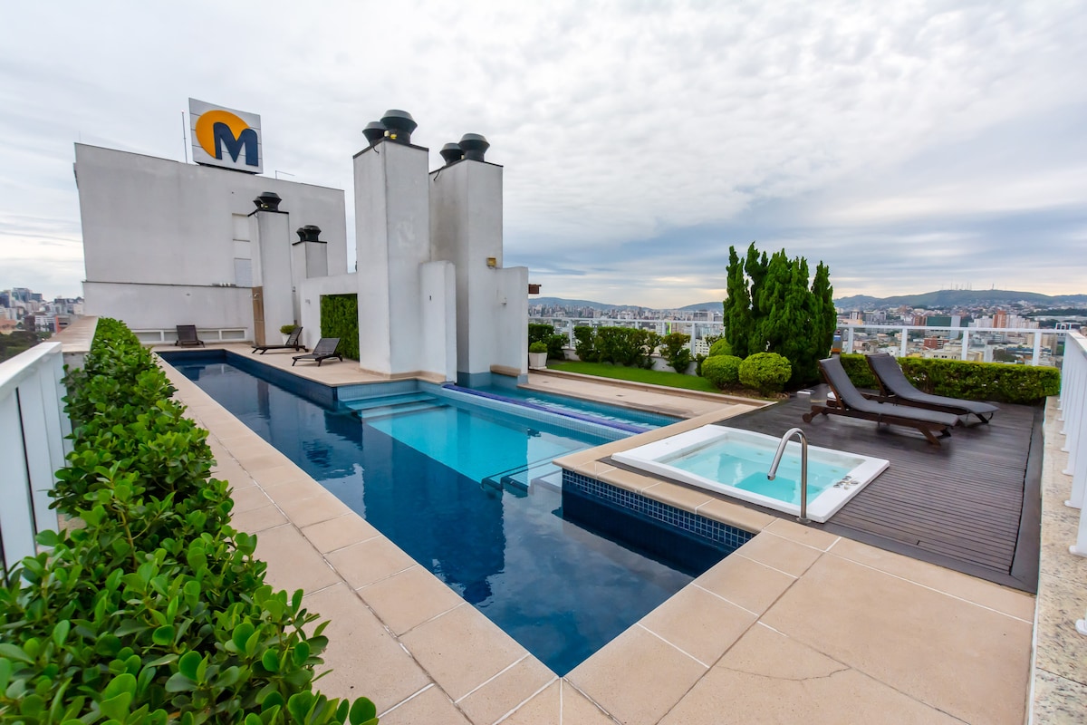 An outdoor area features a rectangular swimming pool and a separate jacuzzi, both surrounded by well-kept greenery. Sun loungers are positioned nearby, offering relaxation options. The view showcases city skyline elements against a cloudy sky, providing a serene urban retreat.