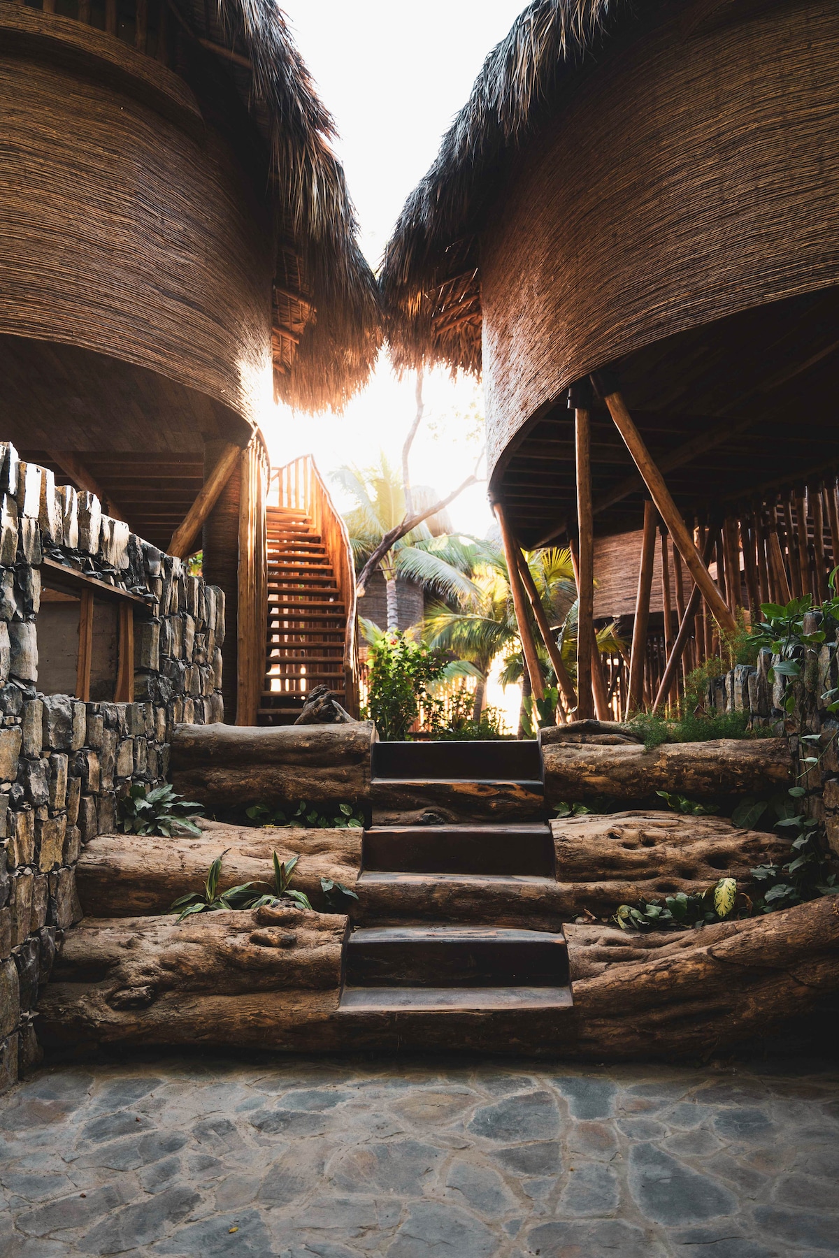 A pathway surrounded by natural stone features guides guests through the space, flanked by two tree houses with thatched roofs. Stairs constructed from natural wood rise toward upper levels, leading to lush tropical vegetation visible in the background.