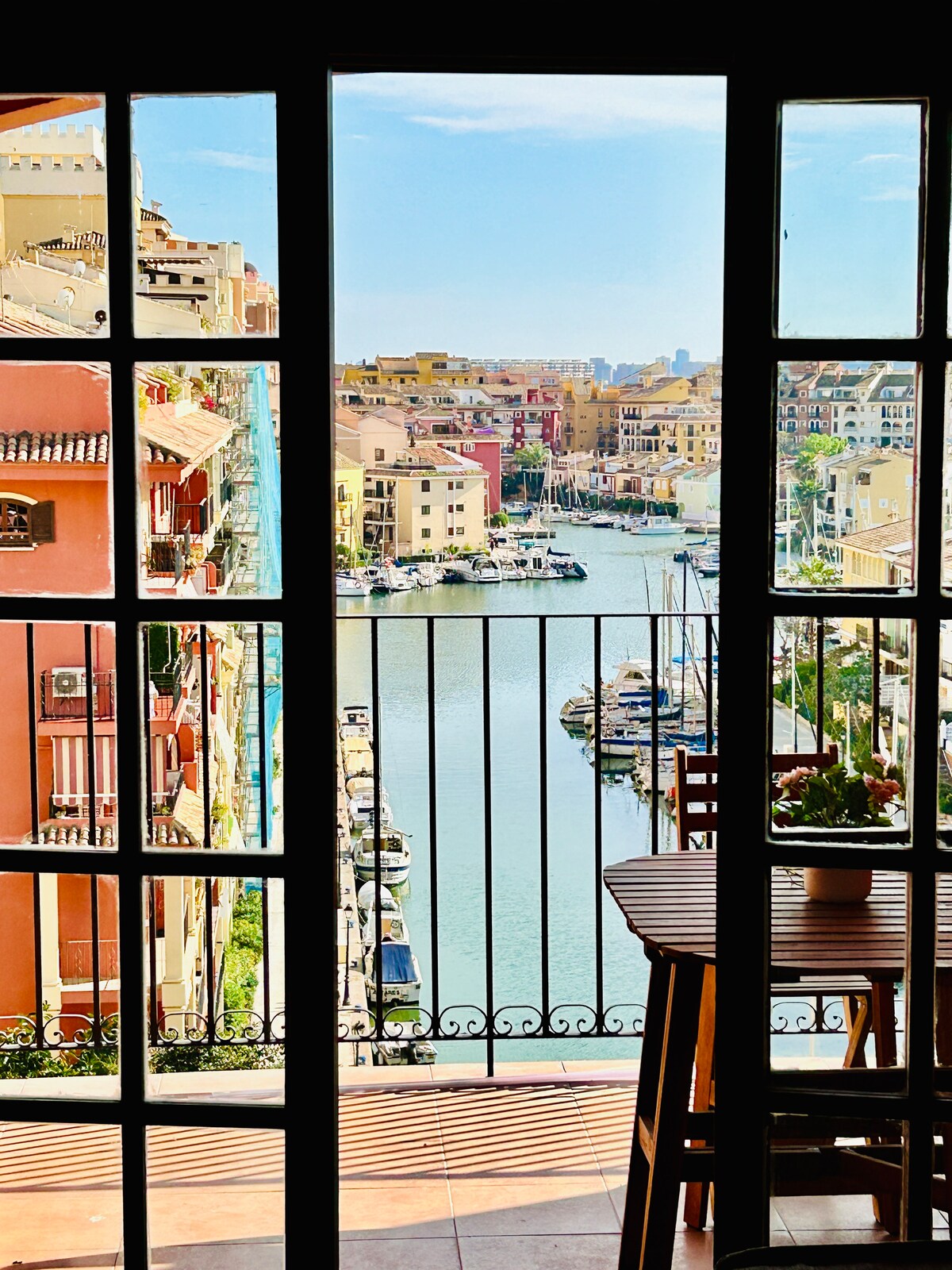 A view of a vibrant waterfront with boats docked along the canal is framed by large glass doors. The scene is complemented by a small dining table and chairs positioned near the entrance, allowing for a picturesque glimpse of the surrounding architecture and clear blue sky.