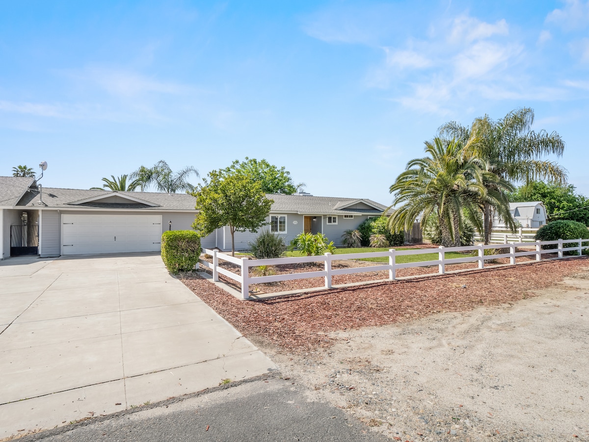 The exterior of a spacious guest house is shown, featuring a well-maintained front yard with lush greenery and palm trees. A white fence borders the property, and a paved driveway leads to a two-car garage. Bright blue skies add to the inviting atmosphere.