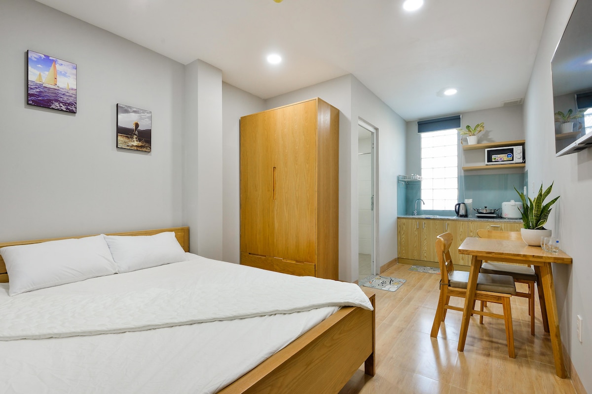 A bright room features a spacious bed with white linens. A wooden wardrobe stands nearby, while a small dining area with a table and two chairs is positioned next to a kitchenette. Natural light comes in through a window, creating a welcoming environment.