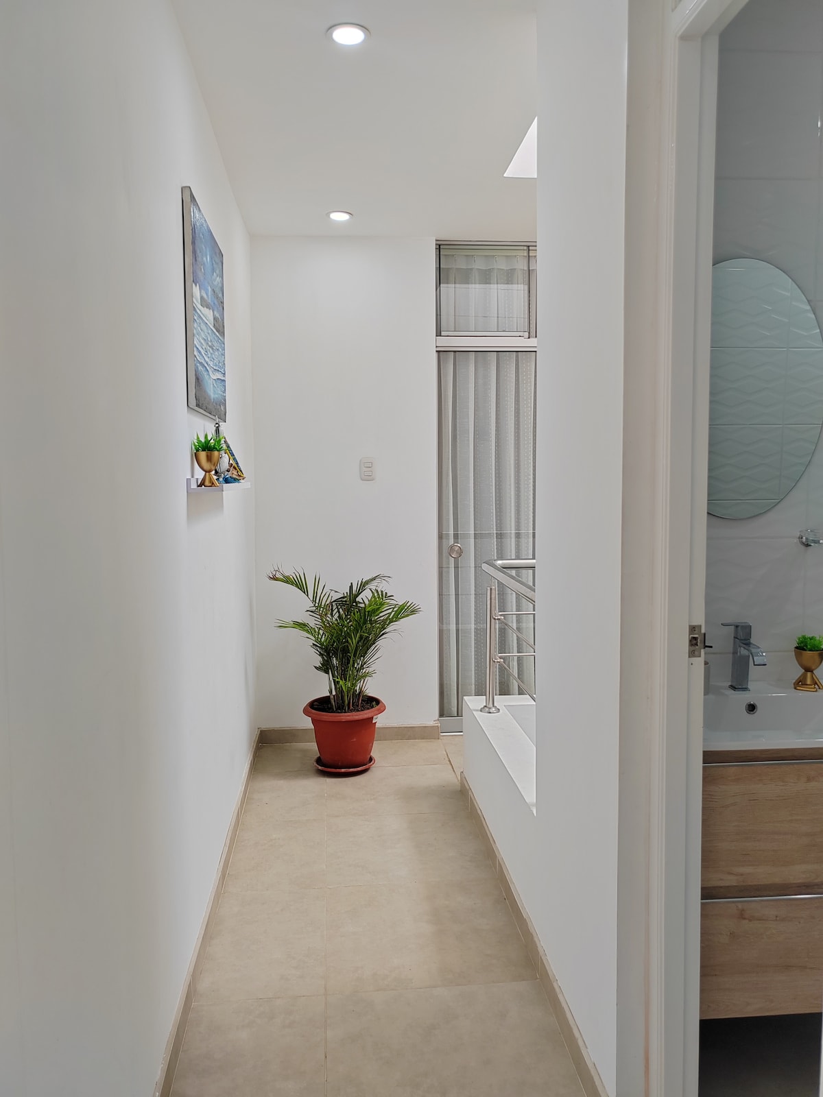 A narrow hallway is lined with light-colored walls, showcasing a small potted plant on the floor. Natural light enters through a window adorned with sheer curtains, creating a bright and airy atmosphere. The hallway leads towards a bathroom area, visible at the end.