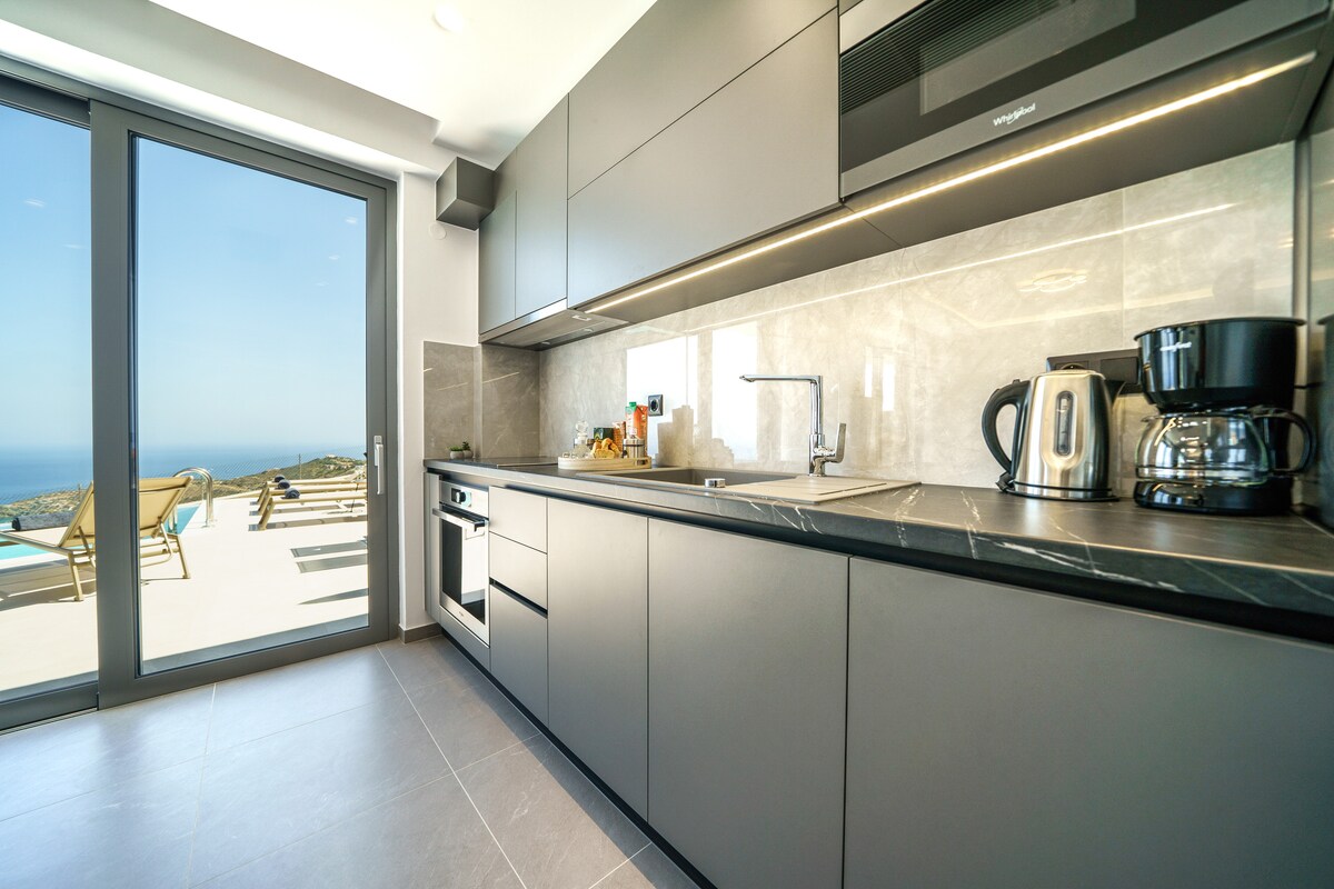 A modern kitchen is highlighted with sleek cabinetry and a spacious countertop. Appliances include a coffee maker and kettle, while scenic views of the sea are visible through large glass doors that open to the outdoor area.