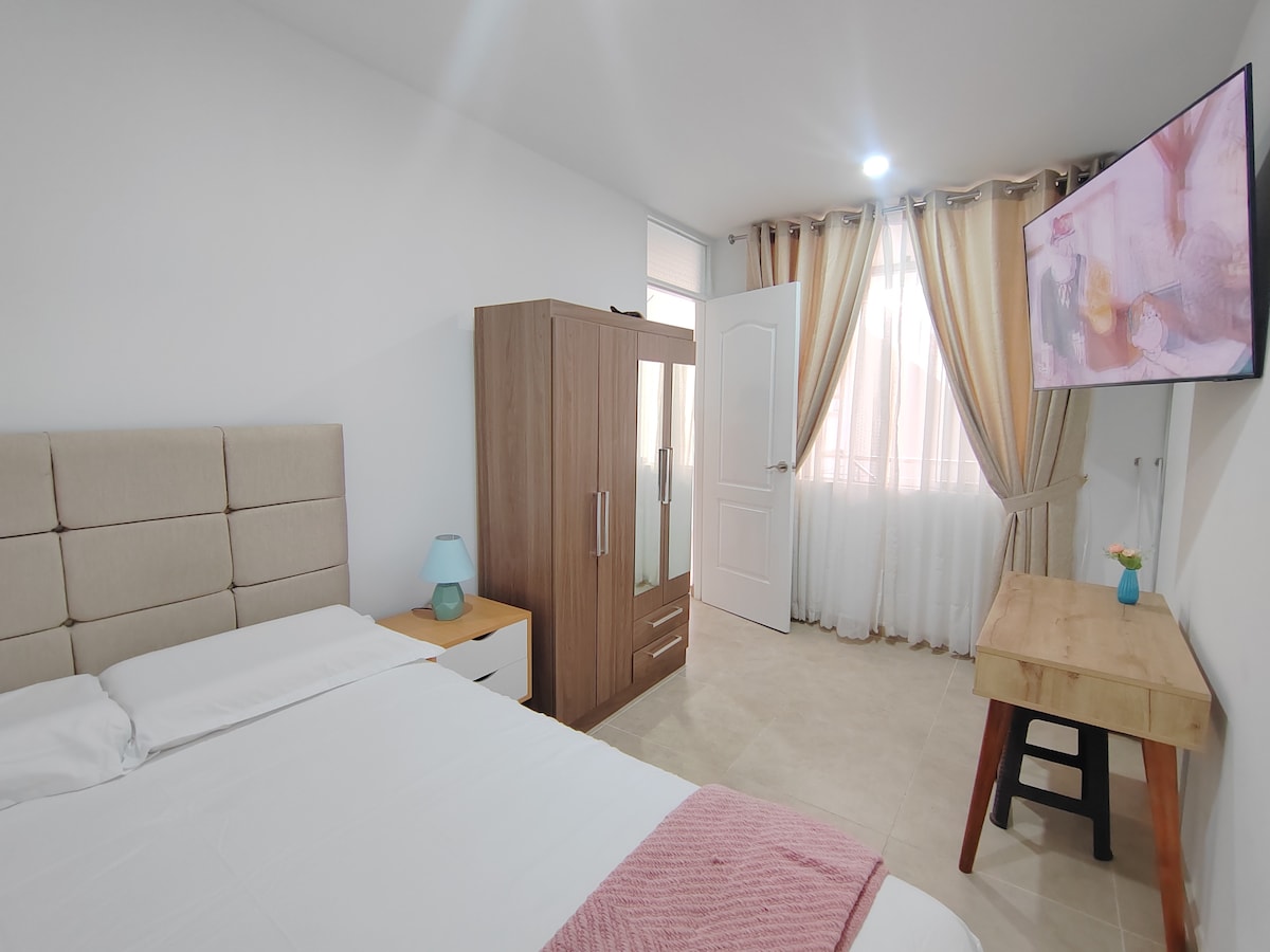 A well-lit bedroom features a spacious bed with light bedding. A wooden wardrobe and bedside table are present, along with a small lamp. Natural light enters through sheer curtains, enhancing the contemporary decor. A wall-mounted television is positioned for viewing.