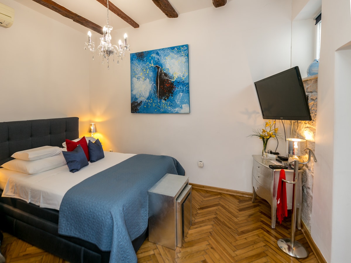 A spacious bedroom is highlighted with wooden beams and an elegant chandelier. A comfortable bed, dressed in crisp white linens and a soft blue coverlet, faces a modern TV. An abstract painting adds a pop of color to the neutral walls, enhancing the inviting ambiance.