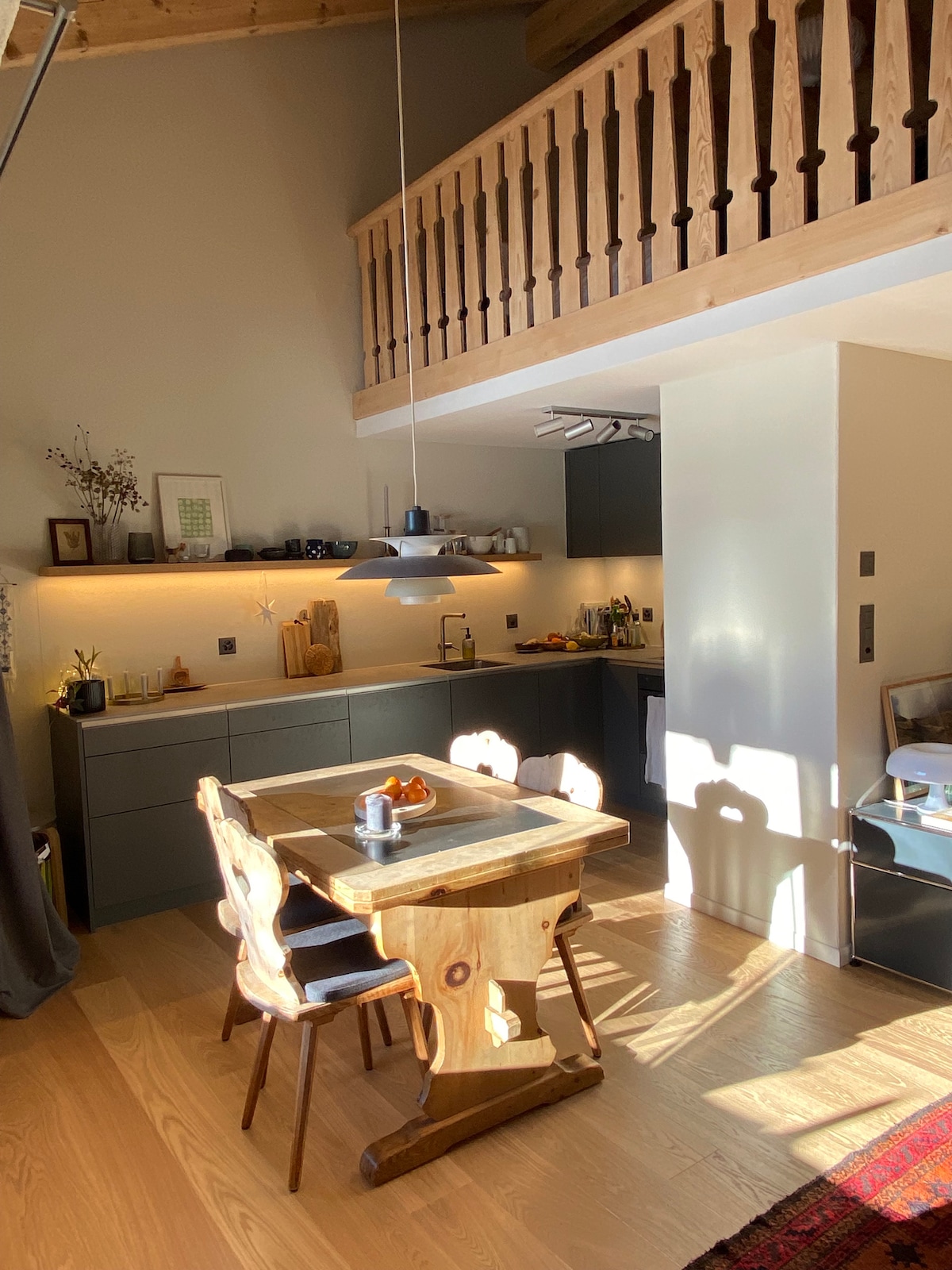 A minimalist kitchen area features dark cabinetry and wooden accents, with a wooden dining table and four chairs. Warm light highlights kitchen utensils, and sunlight casts soft shadows across the floor. A staircase leads to a lofted space visible above.