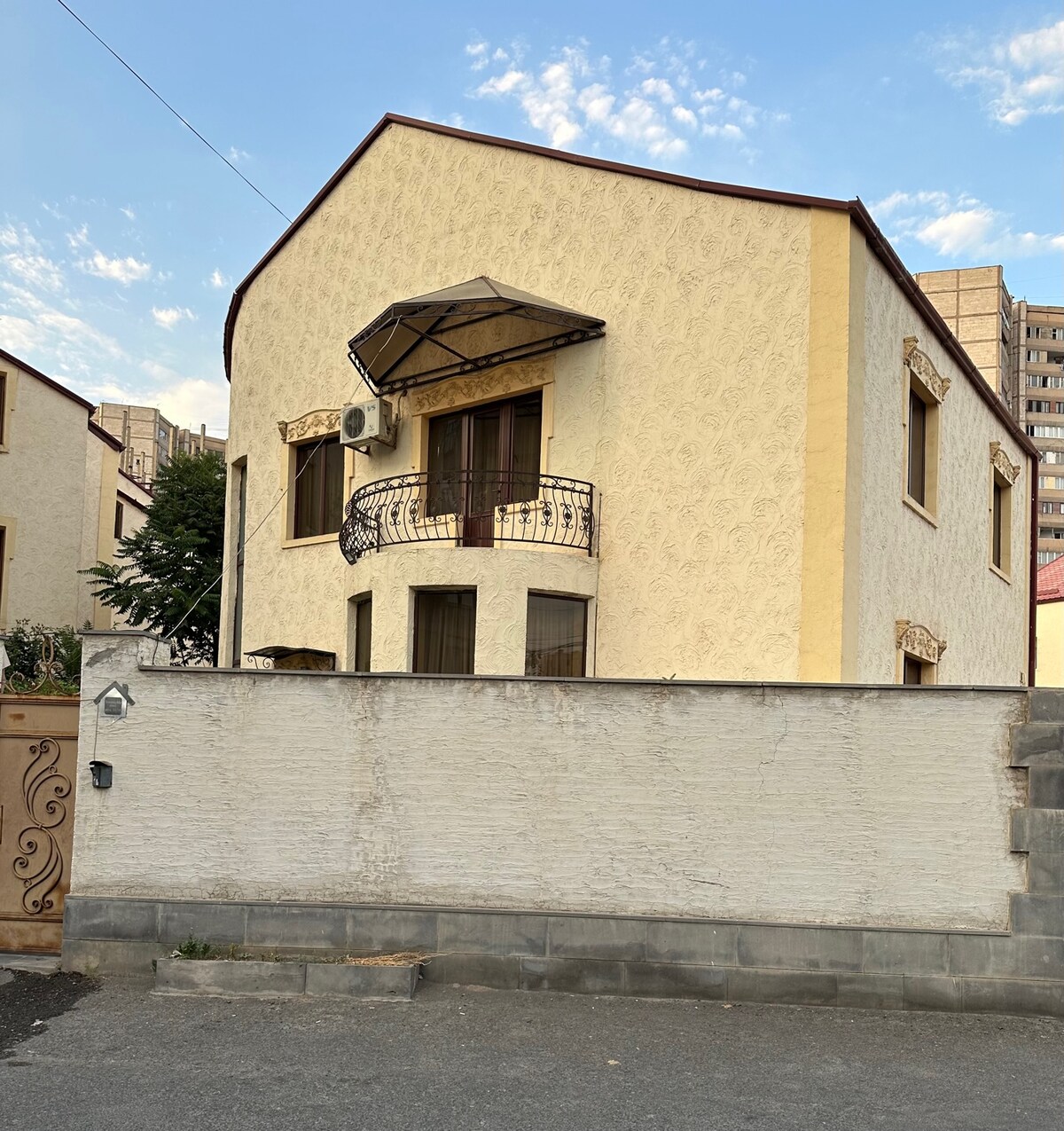 The exterior of a two-story house is presented, showcasing a decorative balcony with ornate railings. The façade, painted in a light color with subtle textures, is complemented by landscaping elements and a surrounding boundary wall. A clear sky is visible above.
