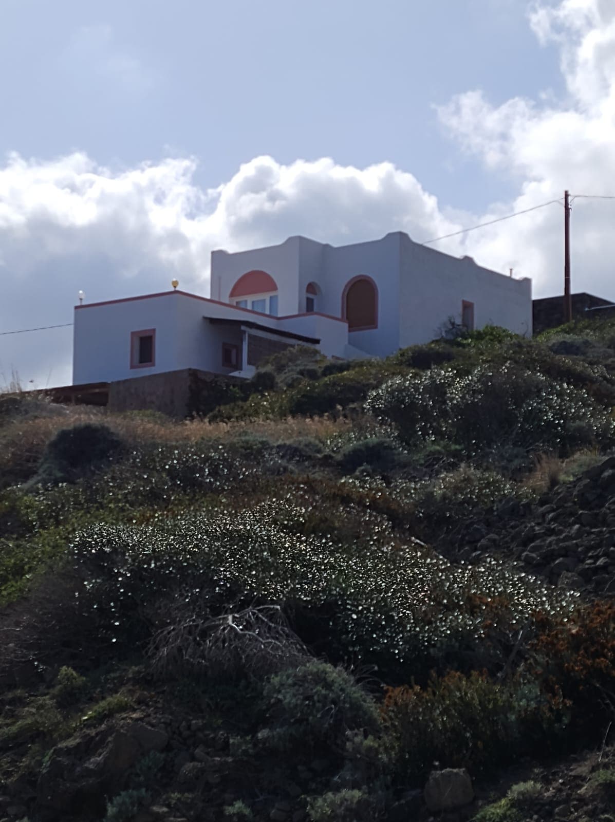 A white-washed house is situated on a sloping terrain, surrounded by greenery and wildflowers. The structure features pink accents around the windows and a circular opening, under a partly cloudy sky. Natural vegetation cascades along the hillside, complementing the tranquil setting.