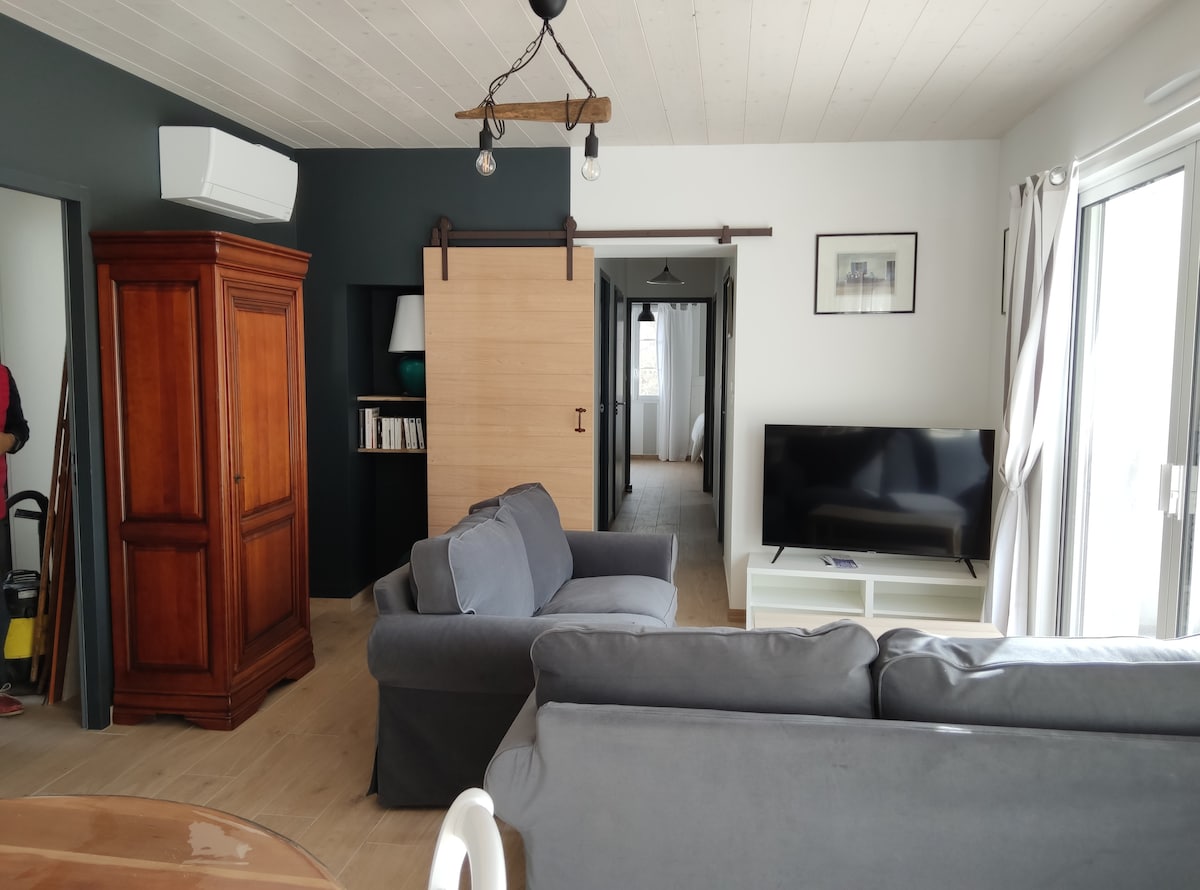 A living area featuring two gray sofas and a sleek television on a white stand. A wooden armoire adds a touch of warmth, while a sliding door leads to a hallway. Natural light enters through the glass doors, enhancing the open and inviting space.