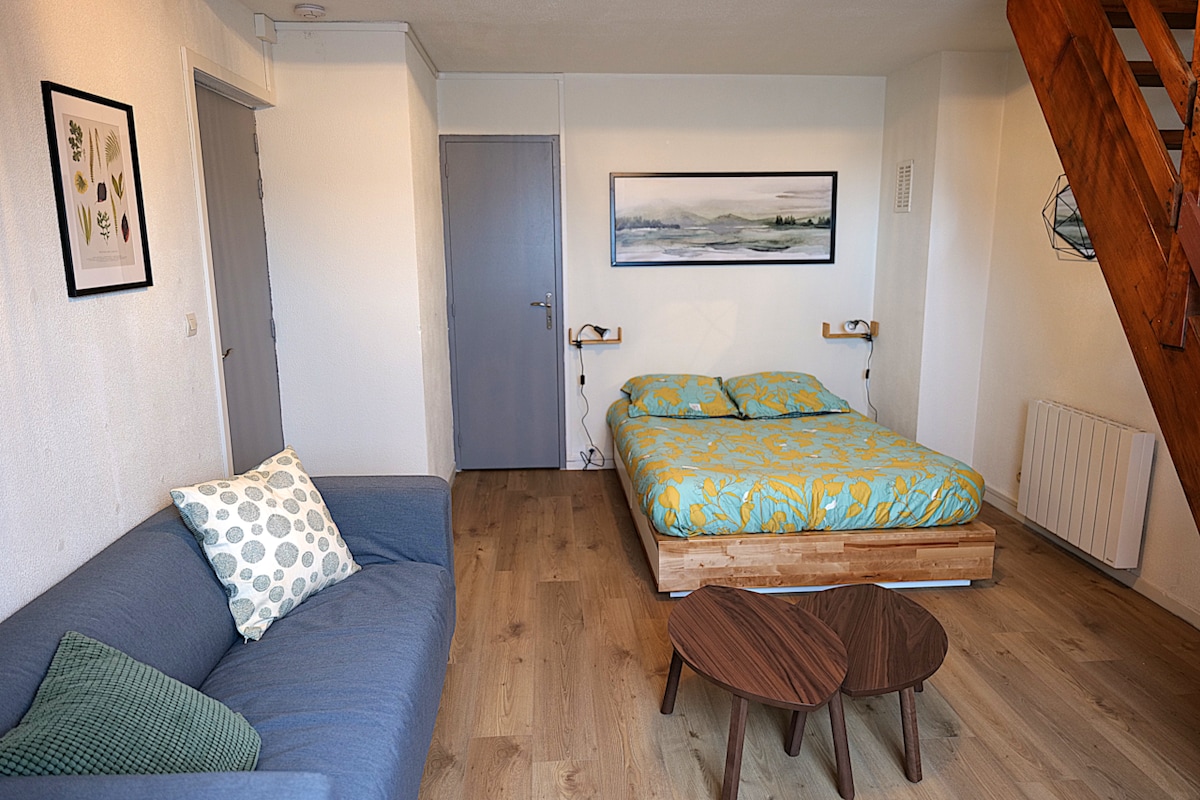 An inviting room features a double bed with a patterned duvet and matching pillows. A cozy sofa is positioned nearby, accompanied by two wooden coffee tables. Light-toned wooden flooring enhances the space, while a piece of nature art adorns the wall above the bed.