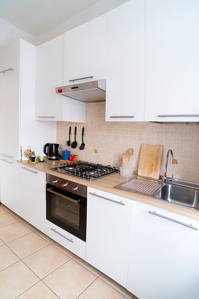 A modern kitchen is featured with a clean, white cabinetry and beige tiled backsplash. Cooking appliances include a gas stove, an oven, and a sink, accompanied by utensils and small kitchen appliances like a kettle. Ample counter space is available for meal preparation.