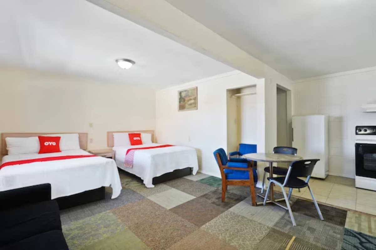 An open living space features two spacious beds with clean, white linens, complemented by a comfortable dark couch. A small dining table with chairs and a kitchenette area, including a refrigerator and stove, are visible. Neutral-toned flooring adds to the inviting layout.