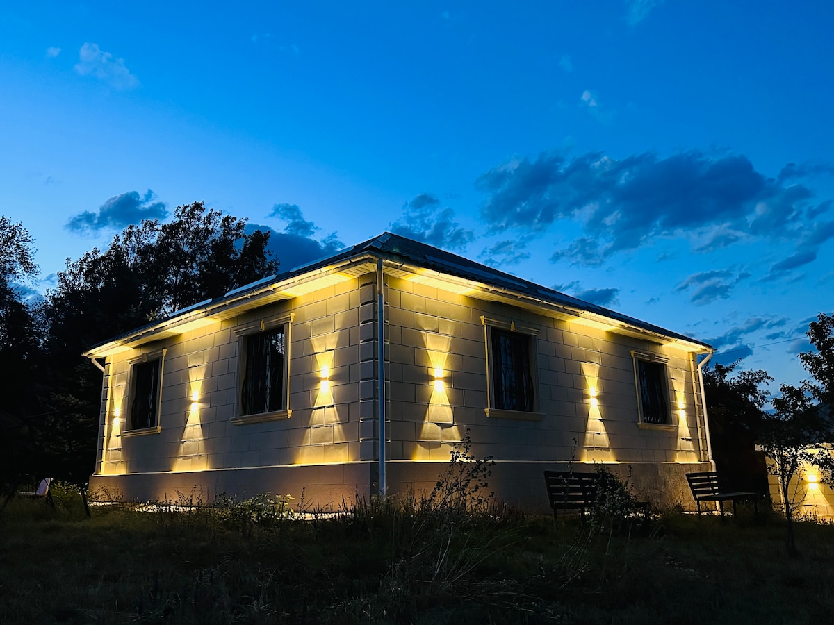 The exterior of a single-story building is illuminated by soft, warm lights, highlighting its textured walls. The evening sky features a blend of blue and gray hues, while grassy areas surround the structure. Several benches are positioned nearby, inviting relaxation.