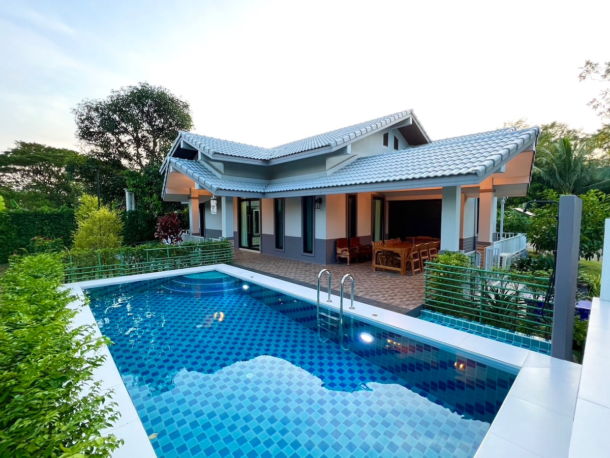A modern pool villa is captured from an elevated angle, featuring a clear blue saltwater pool in the foreground. The outdoor area showcases a tastefully arranged seating area under a shaded roof, surrounded by lush greenery and vibrant landscaping.