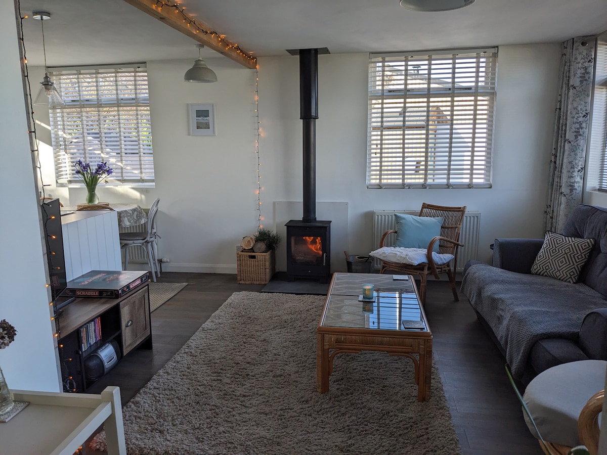 The living room features a comfortable seating area with a sofa and armchair, set against a backdrop of large windows allowing natural light. A coffee table is positioned centrally on a soft area rug, while a wood-burning stove adds warmth to the space.