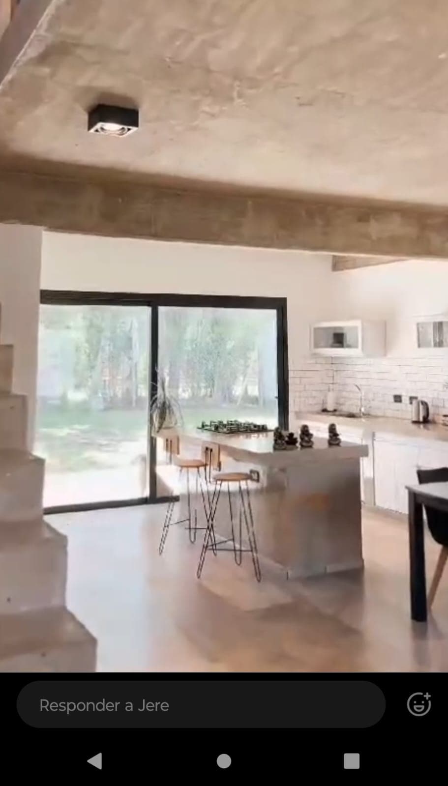 A contemporary kitchen features sleek countertops and a central island equipped with seating. Large glass doors open to a green outdoor space, allowing natural light to fill the area. Modern appliances and white cabinetry complement the minimalist design.