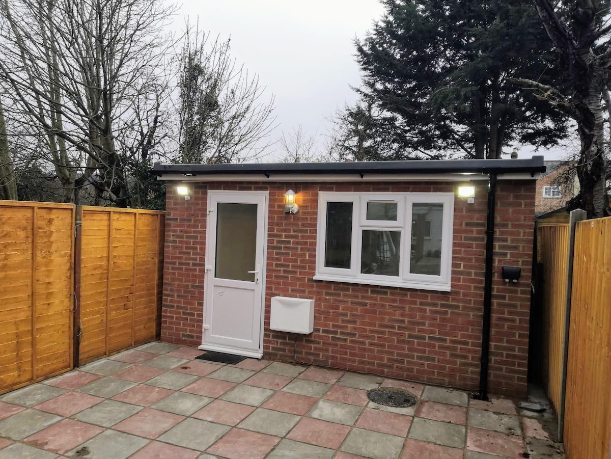 A self-contained entrance to the studio is visible, featuring a white door and a large window. The brick exterior is complemented by wooden fencing on either side, creating a private outdoor space with a paved area. Surrounding trees are present in the background.