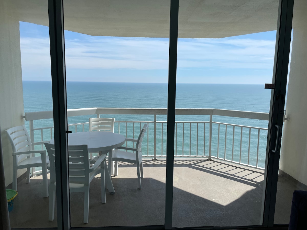 A spacious private balcony is visible, featuring a circular table surrounded by four white chairs. The oceanfront view extends beyond the railing, displaying calm blue waters under a clear sky, creating a serene outdoor space for relaxation.