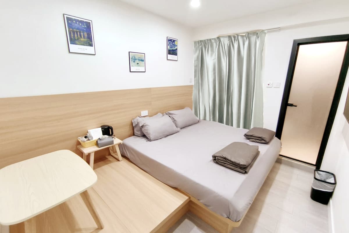 The room features a spacious bed with neutral bedding, accompanied by folded towels. A small table is located beside the bed. Light-colored wooden furnishings are present, and a window is draped with light green curtains, providing a soft ambiance.