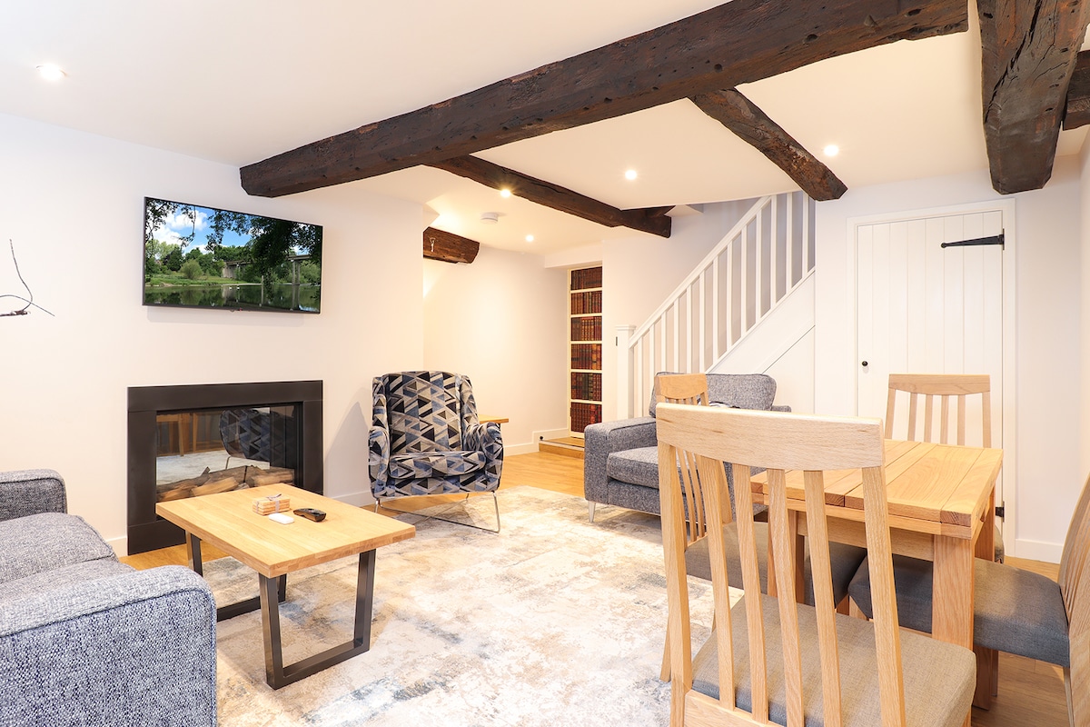 A spacious living area is equipped with a modern television mounted on the wall. Comfortable seating options are arranged around a wooden coffee table, while a dining table with wooden chairs is visible. Exposed wooden beams complement the bright, neutral color palette of the room.