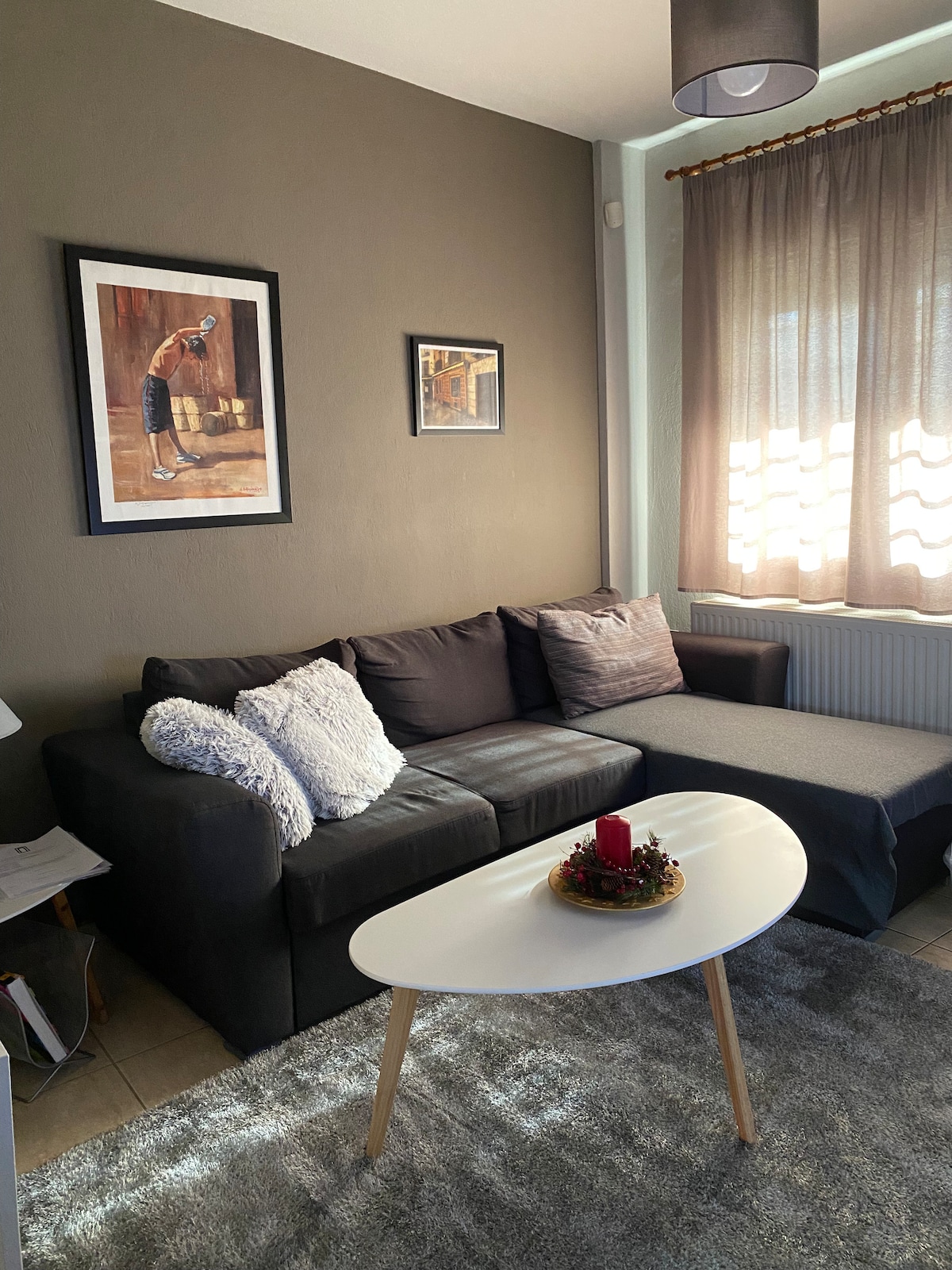 A corner of a living area is shown, featuring a dark sectional sofa with a throw pillow. A small, round coffee table occupies the center space, adorned with a decorative tray. Natural light filters through a partially open window, with soft curtains providing a gentle ambiance.