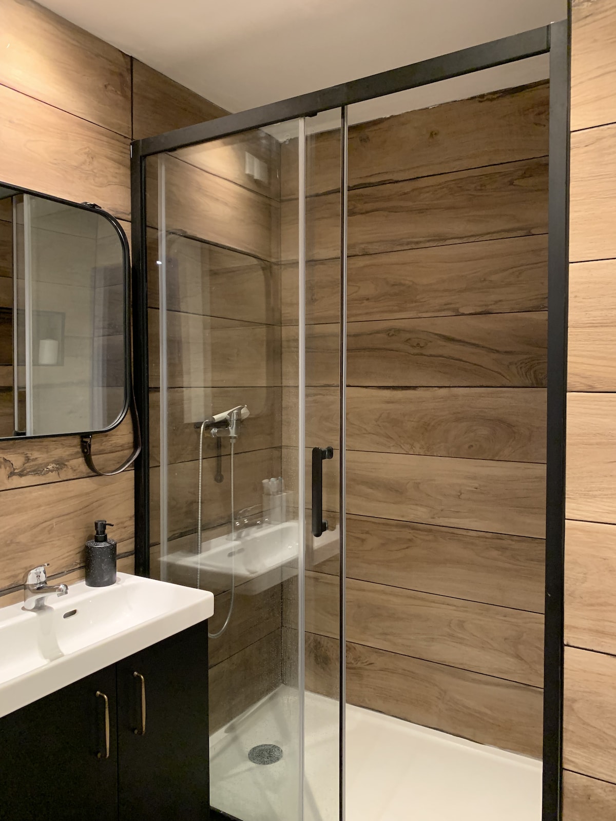 A modern bathroom features a glass shower enclosure with a sleek black frame. Warm wood-like paneling adorns the walls, complementing the minimalist sink vanity below a round mirror. Bathroom essentials are neatly arranged beside the sink, emphasizing a clean and organized space.