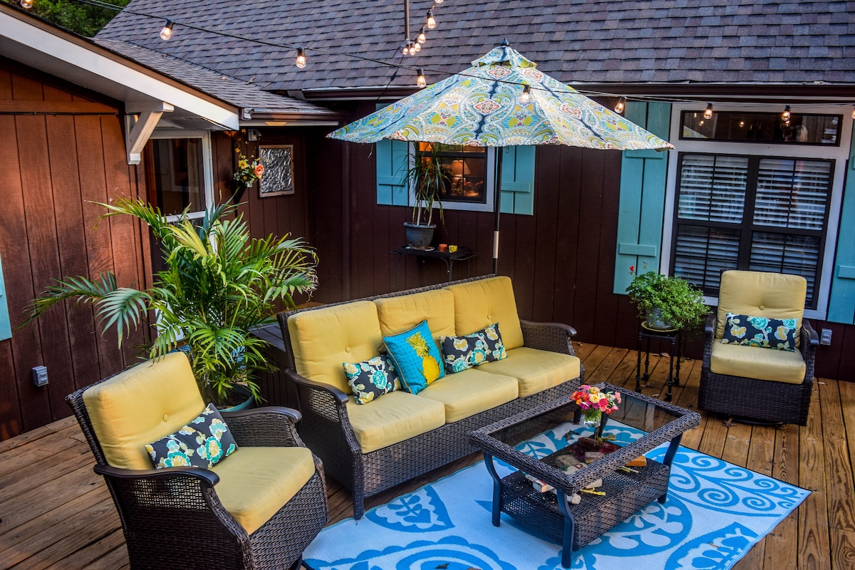 The outdoor seating area features a wicker sofa and two armchairs with vibrant yellow cushions. A decorative umbrella provides shade over a glass coffee table, which is placed on a patterned rug. Potted plants enhance the inviting space along a wood deck, illuminated by string lights.