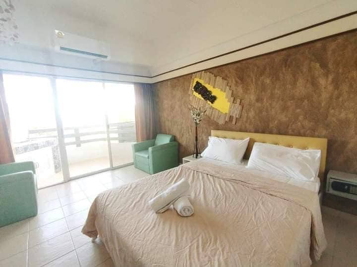 A spacious sleeping area is presented with a large bed, adorned with neutral linens and neatly arranged towels. Two soft green chairs are positioned nearby, and large glass doors provide natural light, opening to a view of the outdoors.