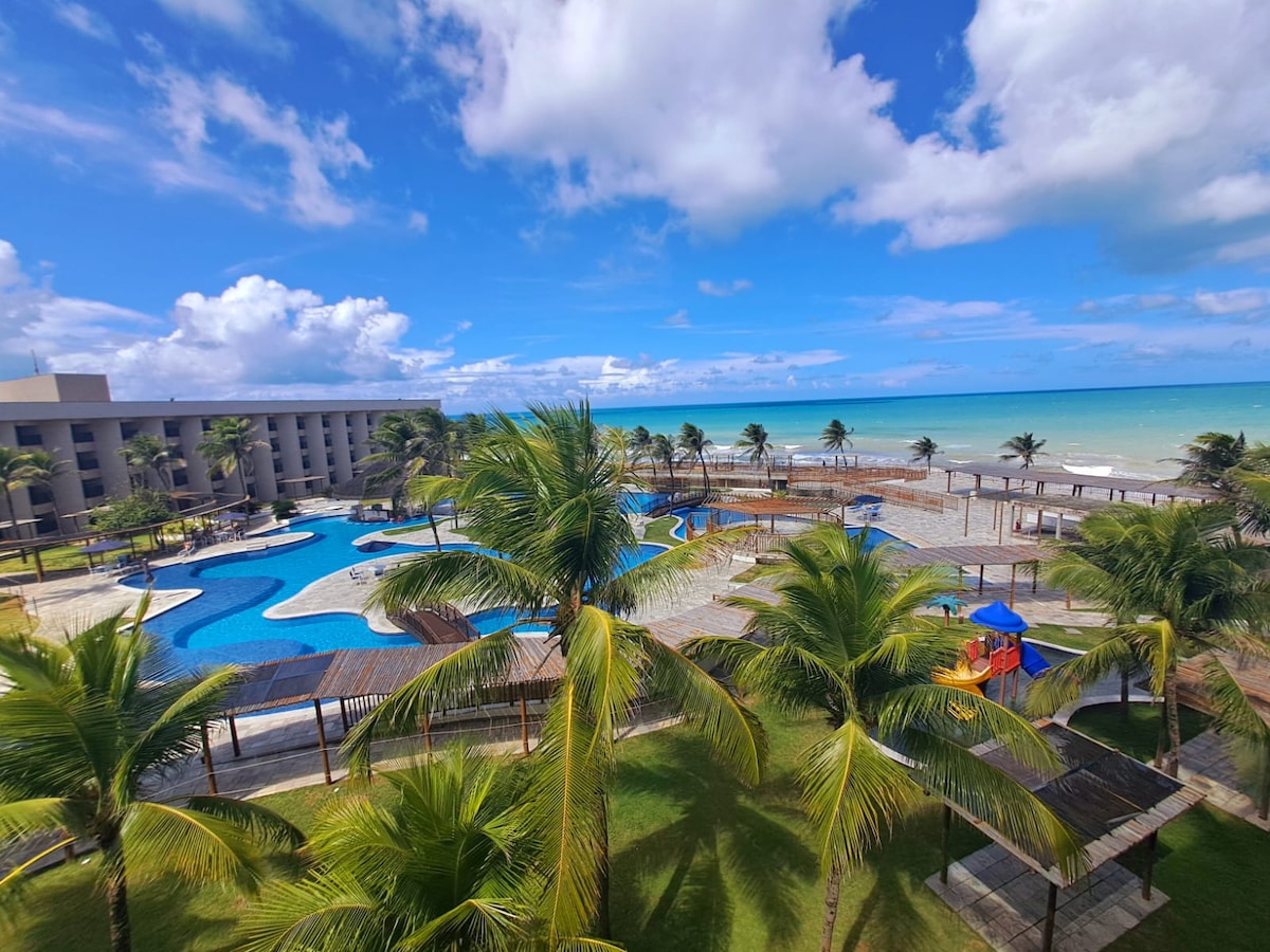 A spacious outdoor area features multiple swimming pools surrounded by palm trees. A private beach is visible in the background, with the ocean meeting a clear blue sky. Lounge areas are positioned throughout the grassy space, providing opportunities for relaxation.