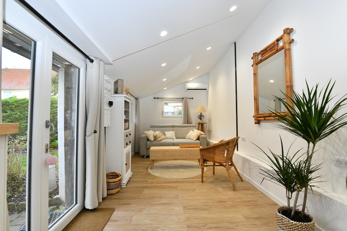 A bright living area features a beige sofa and a textile-covered daybed. A large mirror is mounted on the wall, reflecting the natural light. A potted plant and a wicker chair add warmth to the space. French doors open to a garden view.