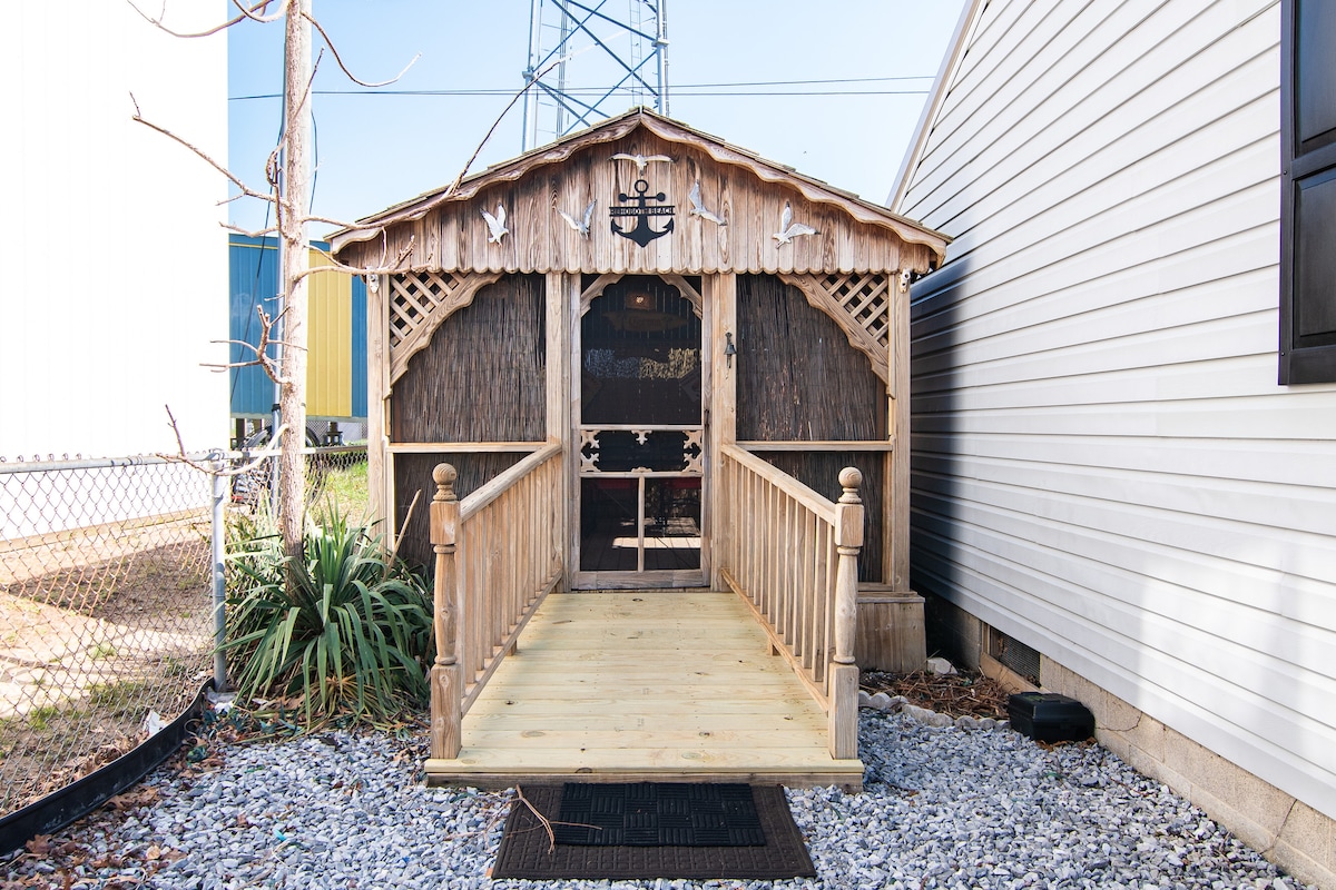 A charming wooden shed features a decorative facade with nautical elements. A wide, level ramp leads to the entrance, framed by minimal landscaping and gravel. The structure is situated between two larger buildings, providing a cozy spot for storing bikes and beach essentials.