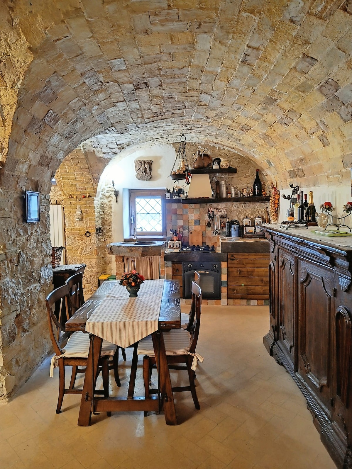 The kitchen showcases a rustic design, featuring an arched stone ceiling and wooden cabinetry. A small dining table with two chairs occupies the space, complemented by a variety of kitchen appliances. Natural light enters through a window, highlighting the warm tones of the stone walls.