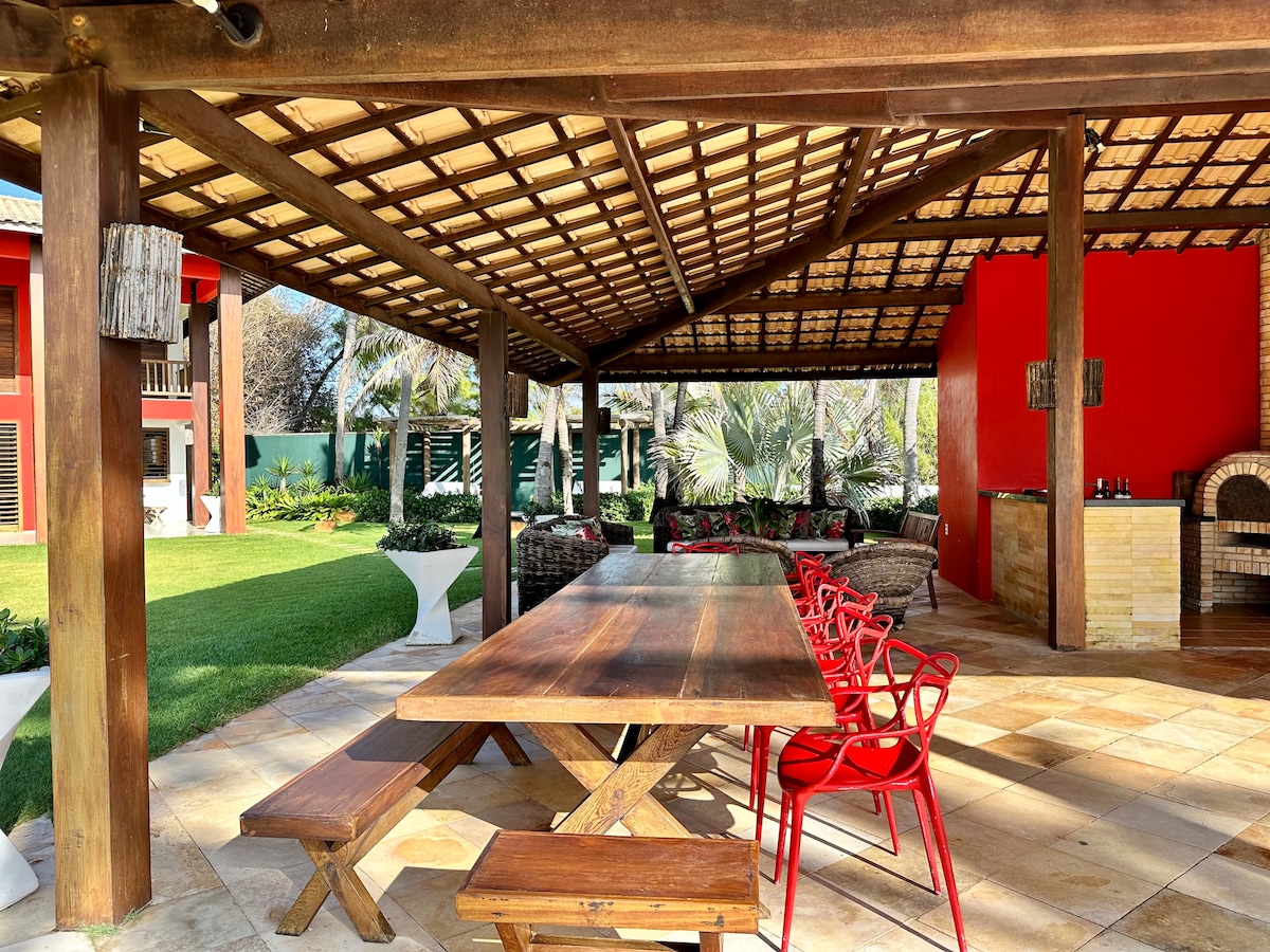 An outdoor dining area features a large wooden table surrounded by bright red chairs. The shaded space is supported by wooden beams and a thatched roof. Lush greenery and a stone barbecue area are visible in the background, creating a welcoming environment for gatherings.