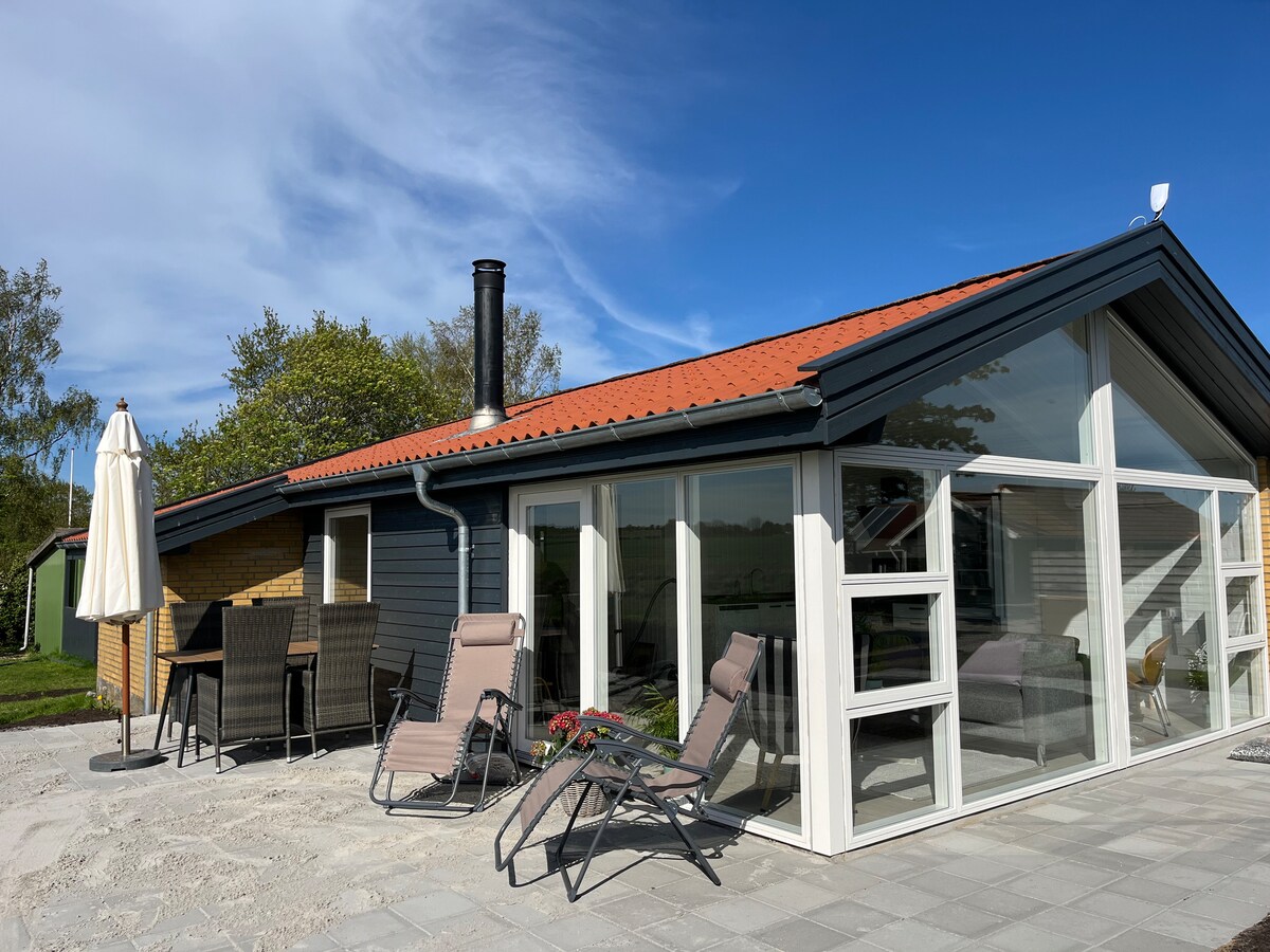 The exterior of the summerhouse is shown, featuring large windows that offer expansive views of the surrounding landscape. A patio area is equipped with comfortable lounge chairs and a dining table. A parasol stands nearby, providing shade on a sunny day.