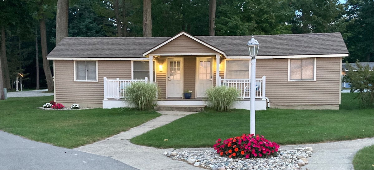 A single-story cabin is set in a green lawn surrounded by trees. Two front doors lead to the entrance, framed by decorative plants. A pathway of steps ascends from the driveway, and a lamp post stands beside vibrant flower beds featuring colorful blooms.